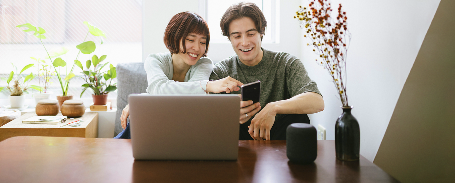 Dos personas frente a una computadora portátil, viendo un teléfono móvil y sonriendo. 