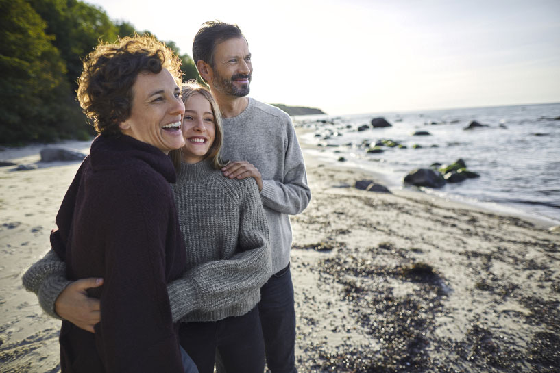 Parents smile and embrace their child as they overlook a rocky ocean shore.